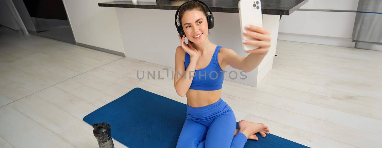 Beautiful and stylish sportswoman, posing for selfie, listens music in wireless headphones, holds smartphone, sits on fitness mat, workout from home.