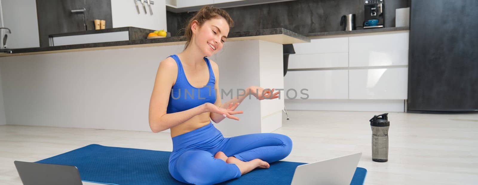 Image of young smiling woman, fitness instructor sitting on yoga mat, talking to client via online video chat on laptop, teaching aerobics, workout training by Benzoix