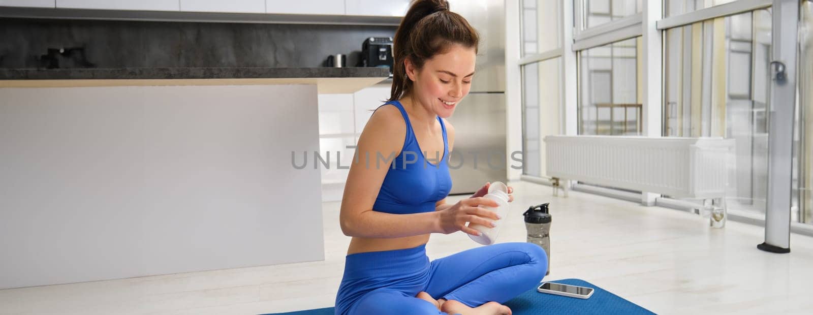 Image of young woman, sporty fitness girl taking vitamins, dietary supplements for muscles strength, sitting on yoga mat and workout.