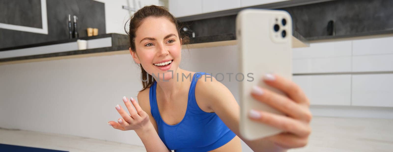 Portrait of young fitness girl, blogger records her workout from home, poses for selfie on smartphone, does yoga on camera, sits in blue sportsbra and leggings by Benzoix