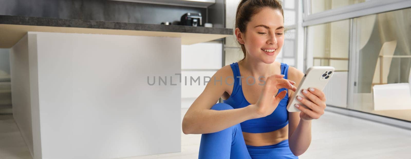 Image of young fit woman wearing sportbra and blue leggings, using smartphone app, does workout from home, sits on floor and follows online training video.