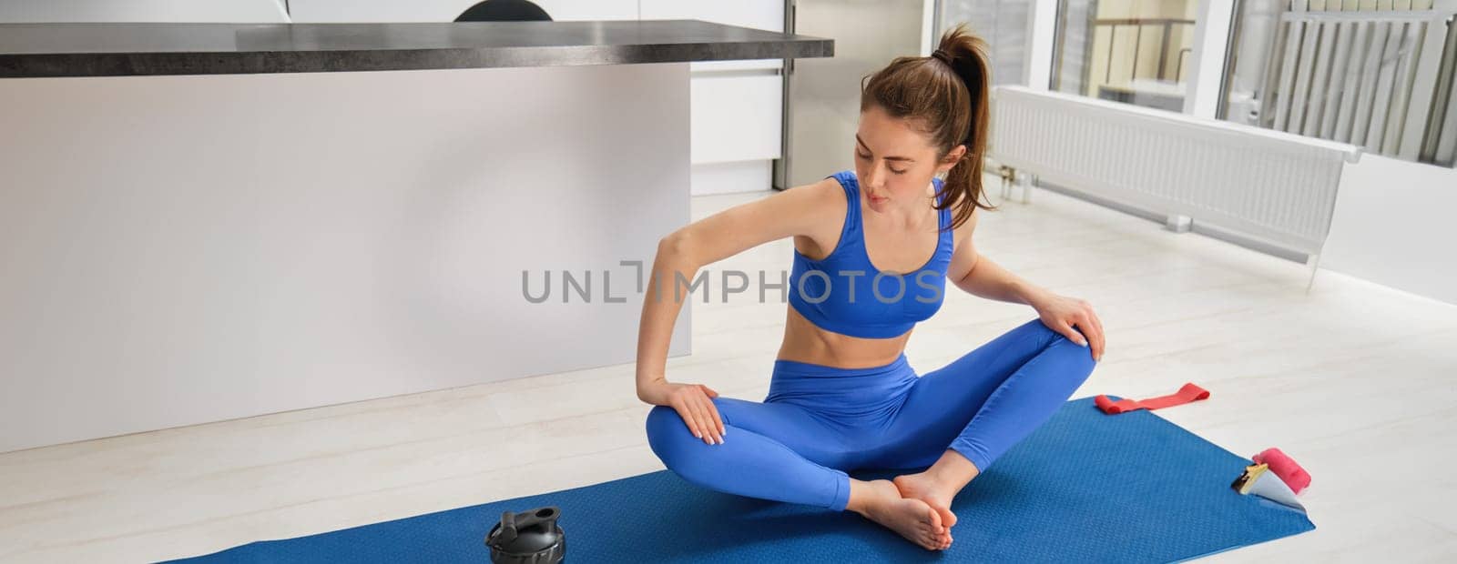 Portrait of young sportswoman, doing sports at home, workout, stretching her legs in lotus pose, smiling while training indoors. Women and lifestyle concept