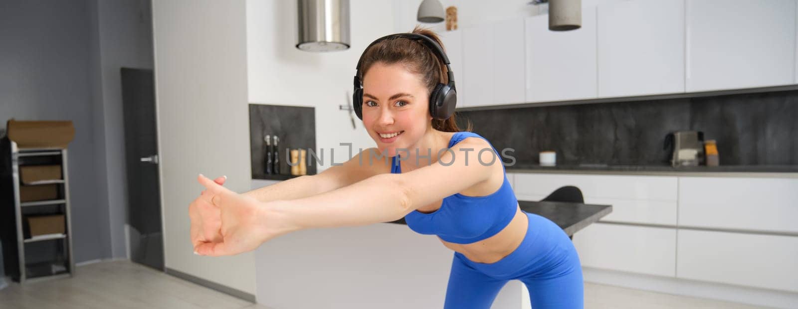 Portrait of beautiful, fit woman in blue leggings and sportsbra, stretching her arms, doing fitness workout, aerobics exercises at home, listening music in wireless headphones by Benzoix