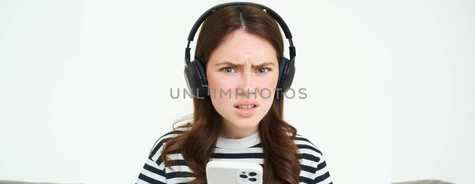 Image of puzzled young woman in headphones, holds smartphone, frowning and looking confused at camera, standing over white background by Benzoix