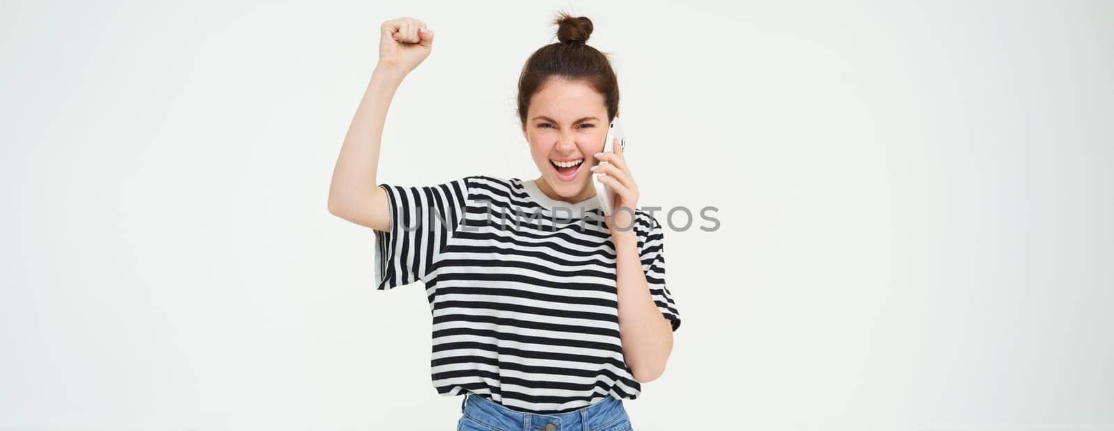 Enthusiastic girl chanting, answers phone call and looks excited, triumphing over great news, stands over white background by Benzoix