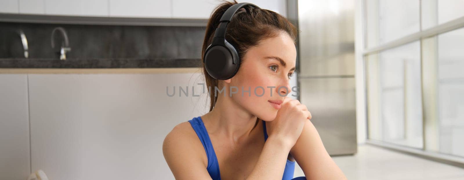Close up portrait of beautiful brunette woman, wearing headphones, listening to music and working out, smiling and looking aside thoughtful. Wellbeing and lifestyle concept
