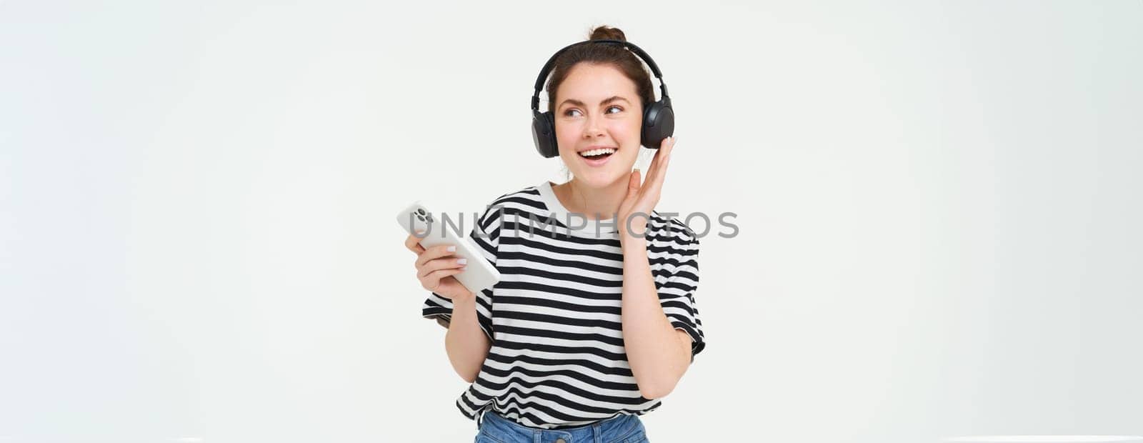 Portrait of happy woman with smartphone changes song on mobile phone streaming app, listens music in headphones, dancing against white background.