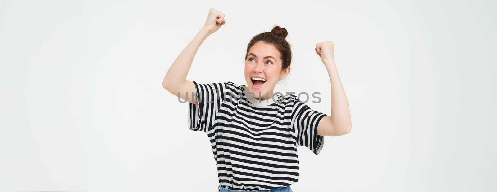 Image of excited young girl cheering, raising hands up, screams from excitement and happiness, winning, celebrating victory, standing over white background.