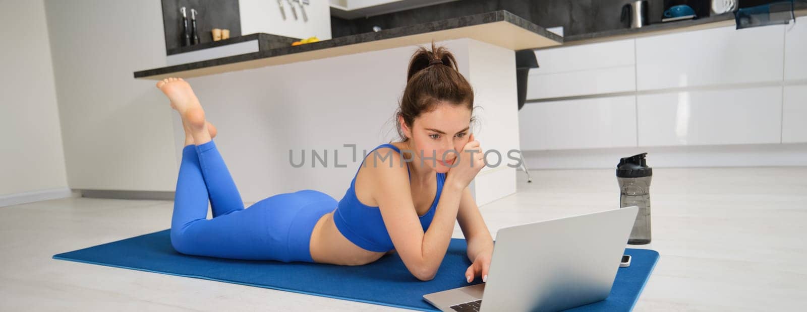 Image of fitness girl laying on yoga mat at home, watching video lesson workout on laptop.
