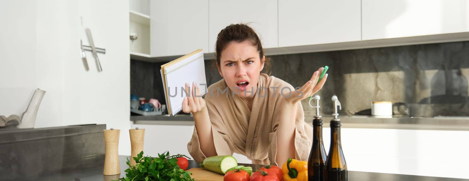 Portrait of woman with angry face, standing near vegetables and looking frustrated, holding notebook, annoyed while cooking meal by Benzoix