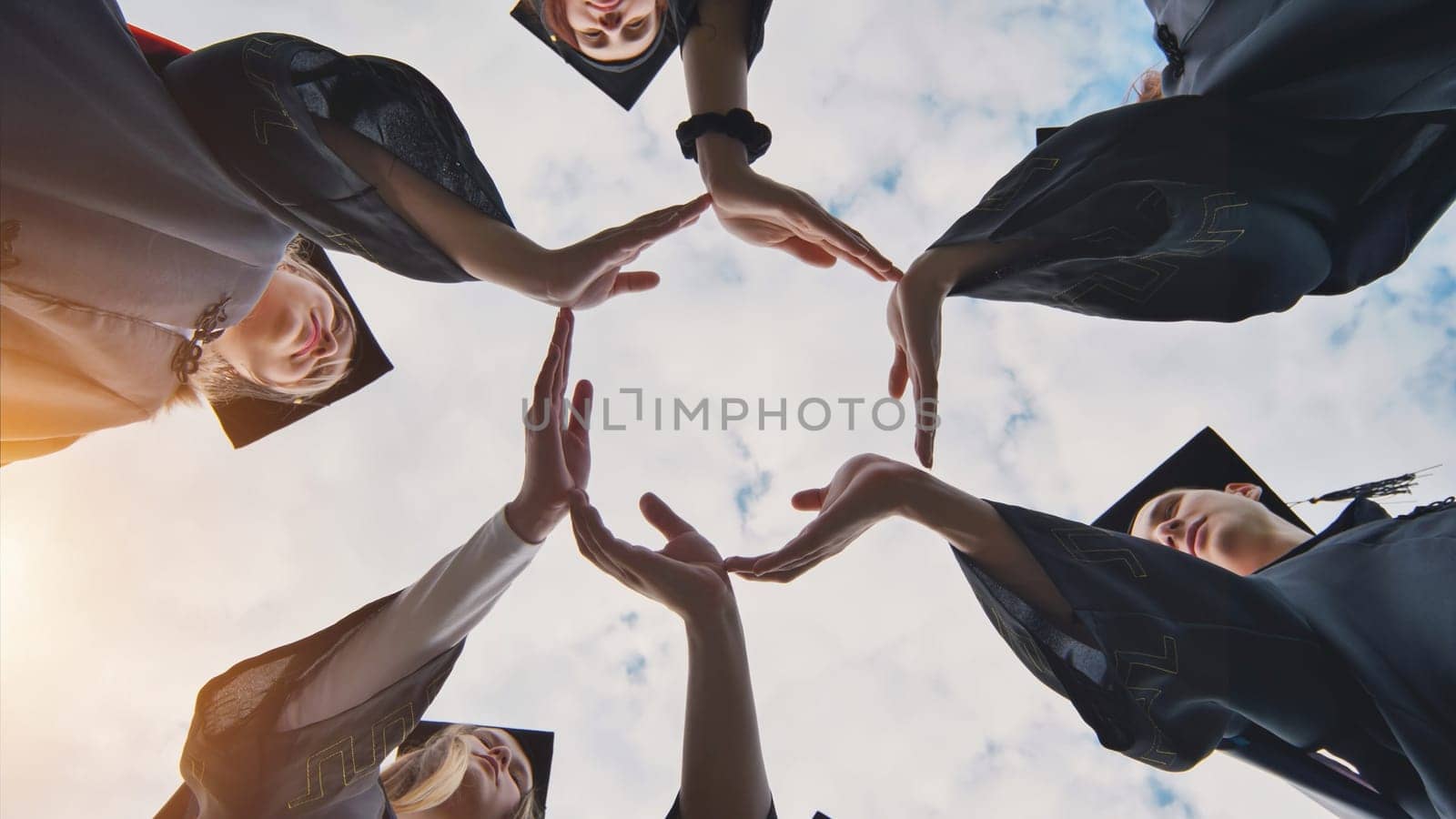 Graduate students in black robes and caps make a circle shape from their hands. by DovidPro