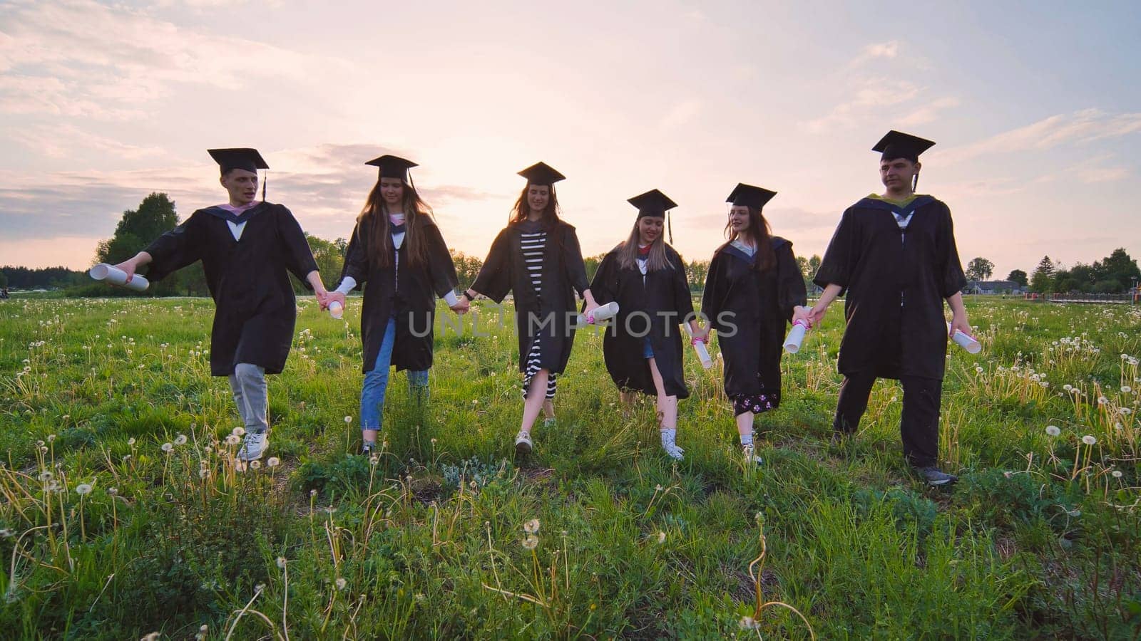 Six graduates in robes walking through a meadow in the evening