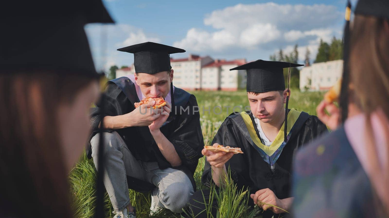 Graduates in black suits eating pizza in a city meadow