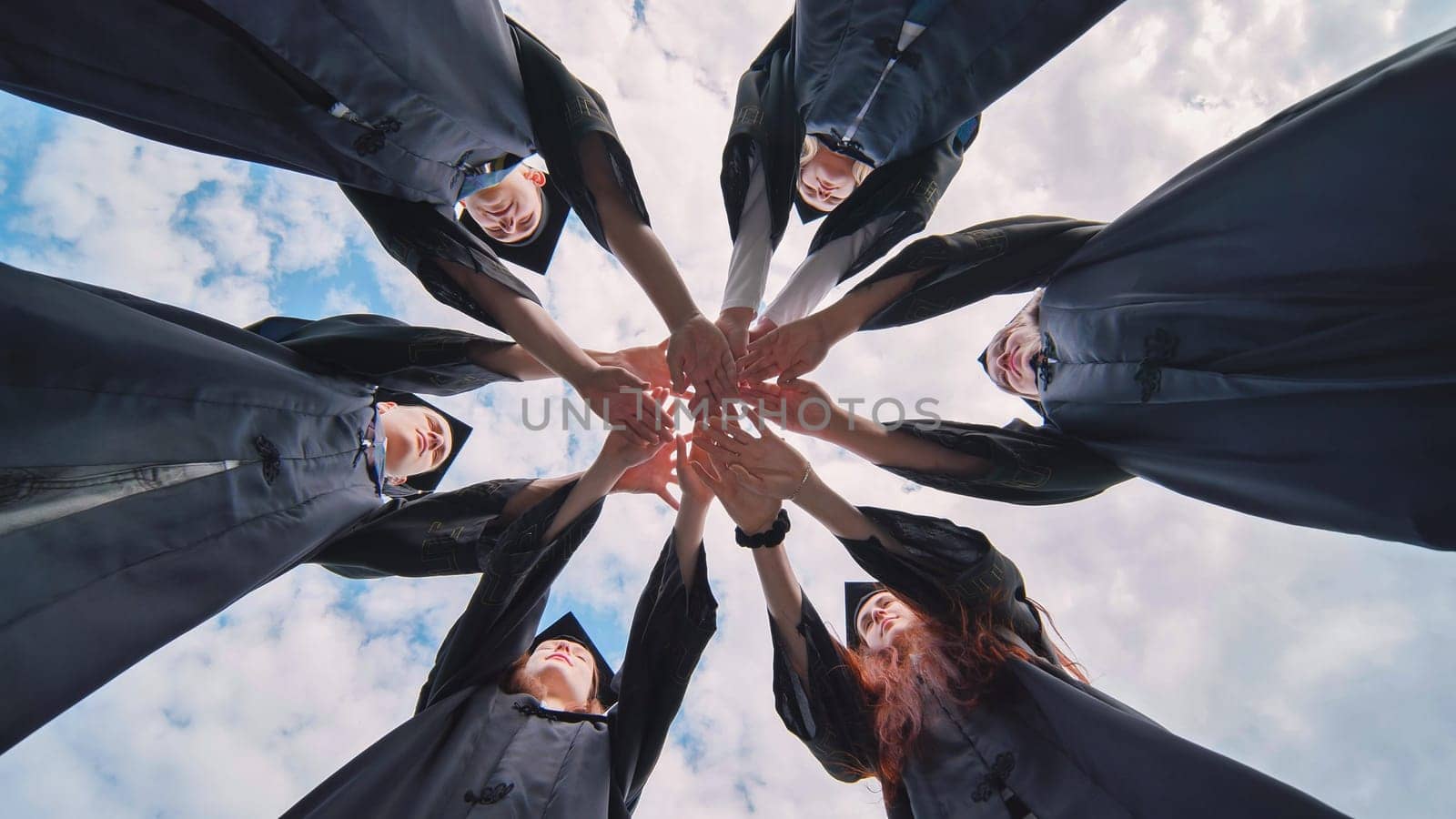 Team of college or university students celebrating graduation. Group of happy successful graduates in academic hats and robes standing in circle and putting their hands together