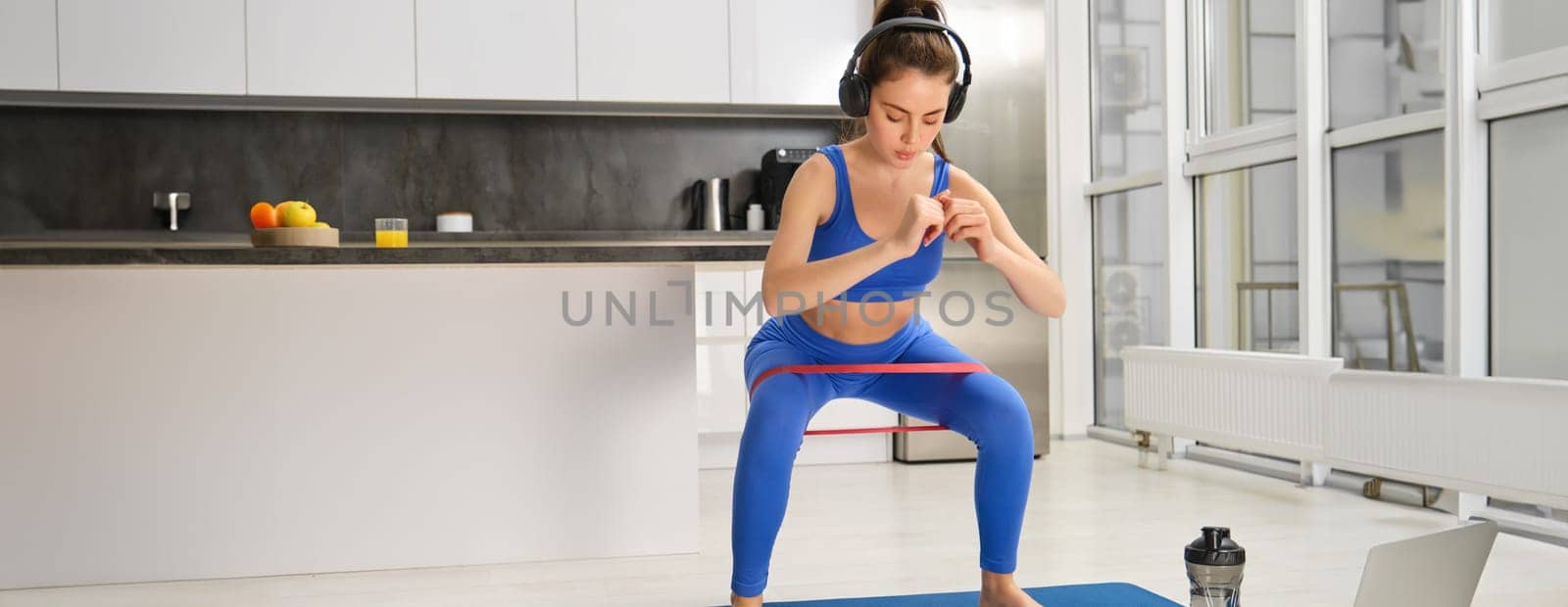 Portrait of active and healthy young woman, doing squats with resistance band, workout at home, listening music in wireless headphones.