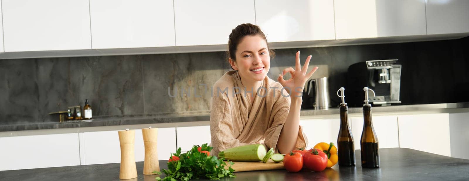 Portrait of smiling brunette girl making vegan dinner, showing okay sign, recommending healthy food, chopping vegetables in kitchen for salad or for vegan breakfast by Benzoix