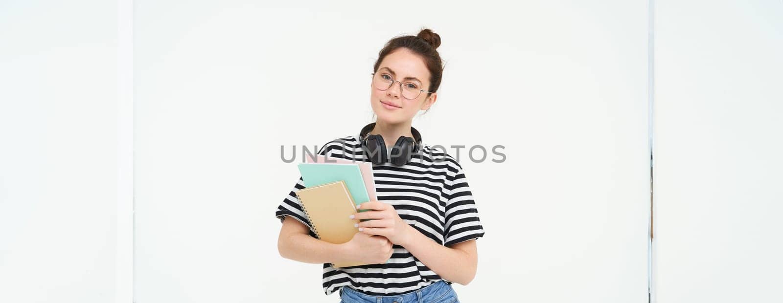 Student and education concept. Young woman with books, notes and pen standing over white background, college girl with headphones over neck posing in studio.