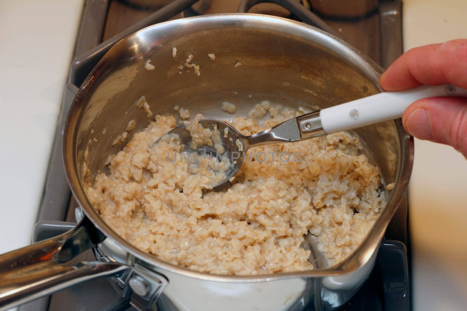 Hand with a tablespoon stirring just cooked oatmeal in a stainless steel pan on the burner of a gas stove cook top.