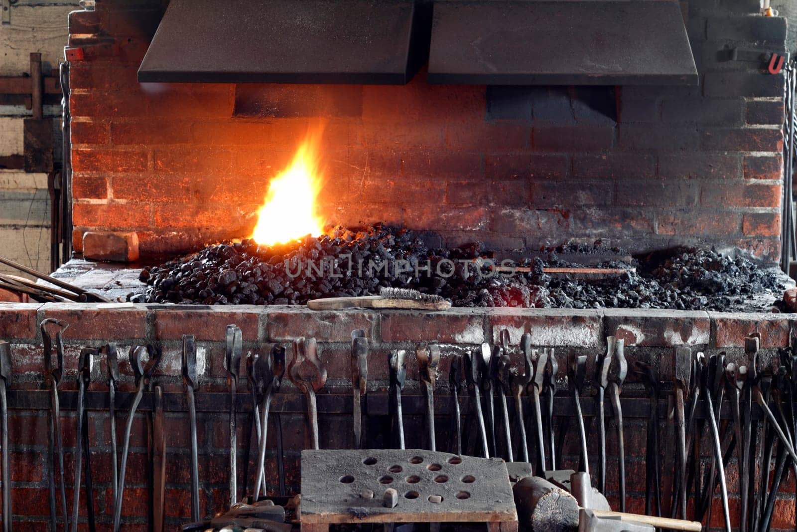 Historic brick blacksmith stove with blacksmith tools in front and a fire burning from coals on top under vents.