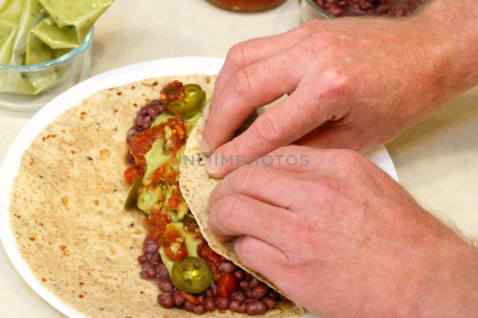 Two hands rolling up in a soft multi grain tortilla adzuki beans, salsa, guacamole, and tamed jalapenos for lunch.