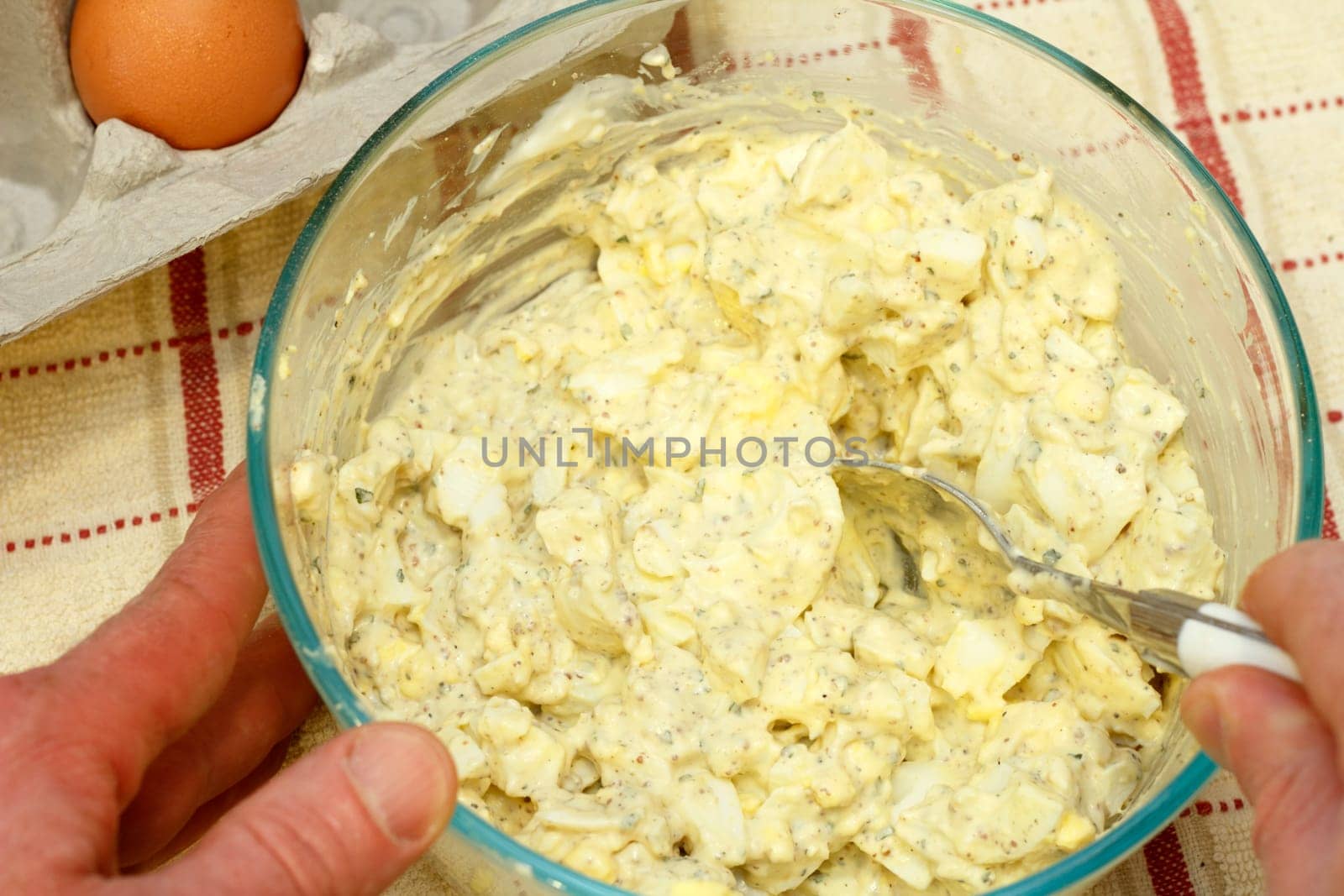 Person mixing egg salad recipe in a glass bowl next to an organic free range brown egg in a cardboard carton on the counter under a tan and red kitchen towel.