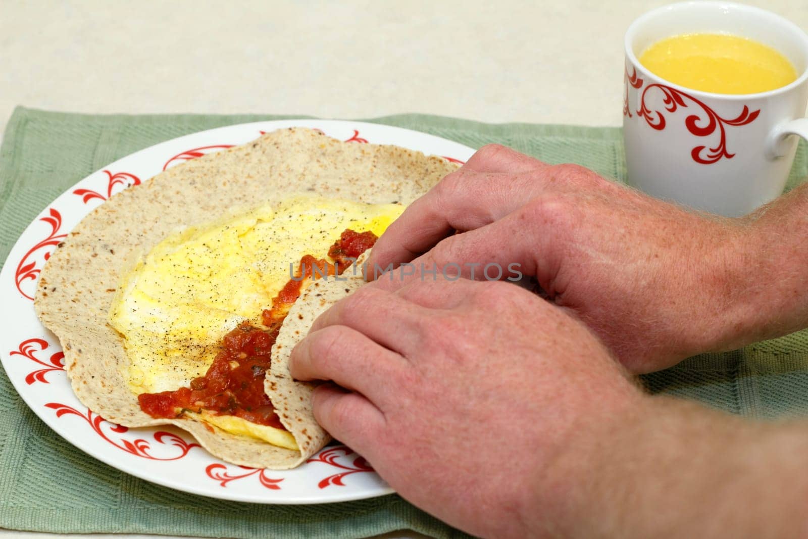 Two hands wrapping up two cooked eggs, tomato salsa, and black pepper in a multi grain soft tortilla with a cup of orange juice on the side.