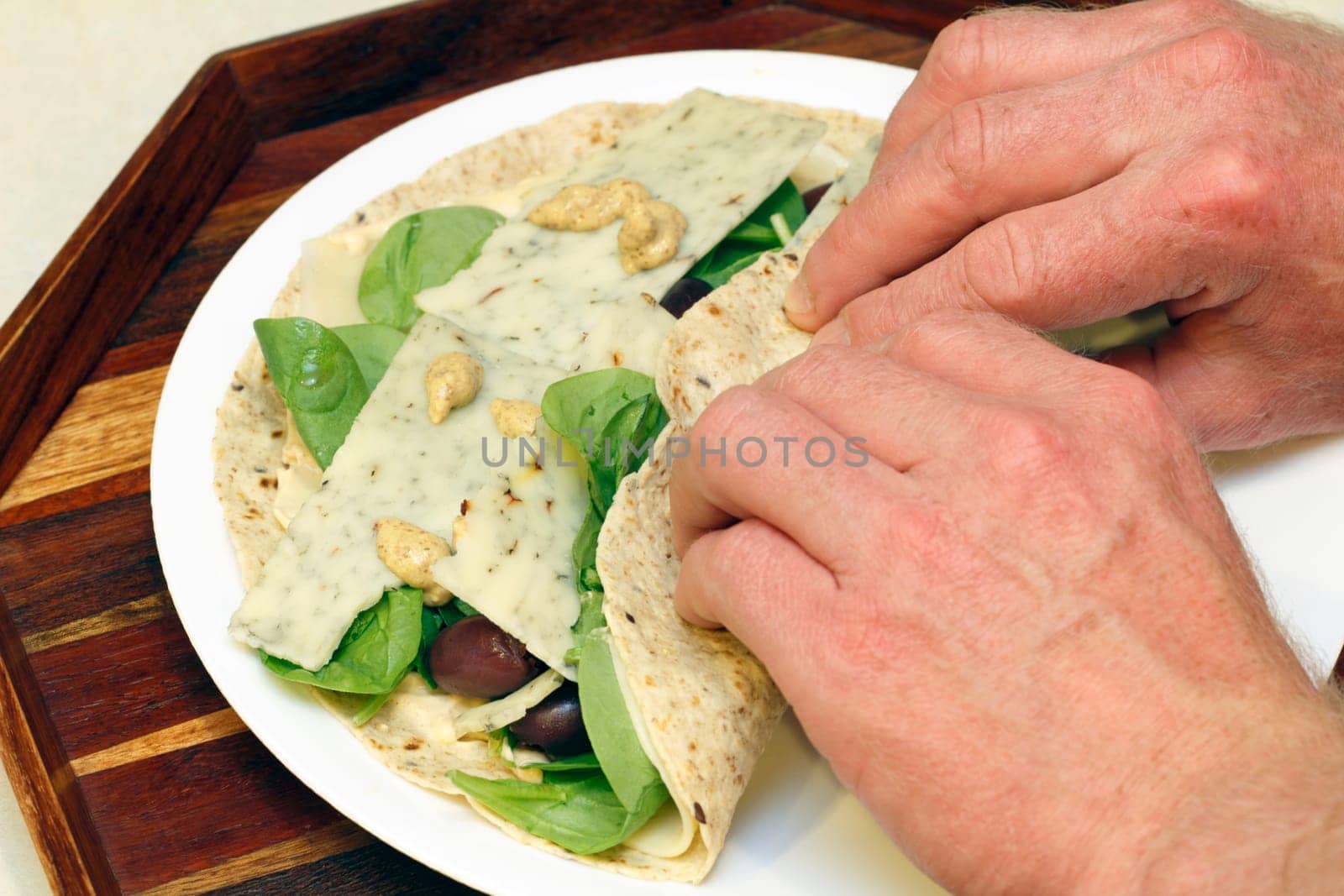 Hands wrapping flavored cheese, spinach, kalamata olives and mustard in a soft multi grain tortilla for lunch.