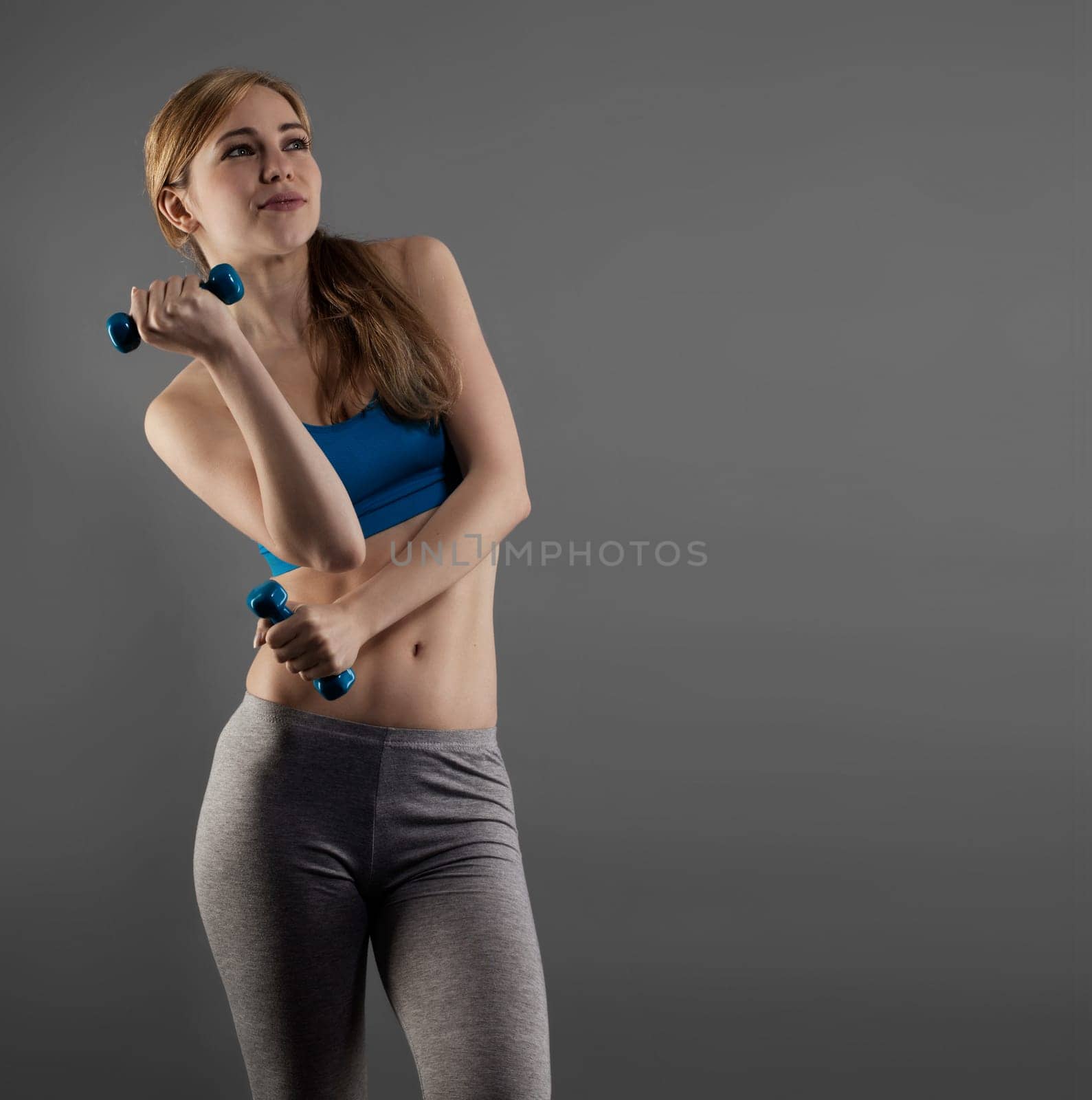 Young sportswoman with fitness dumbbells against gray background