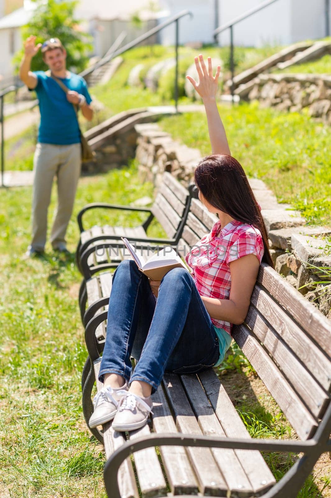 Woman waving to man sitting on bench holding book happy