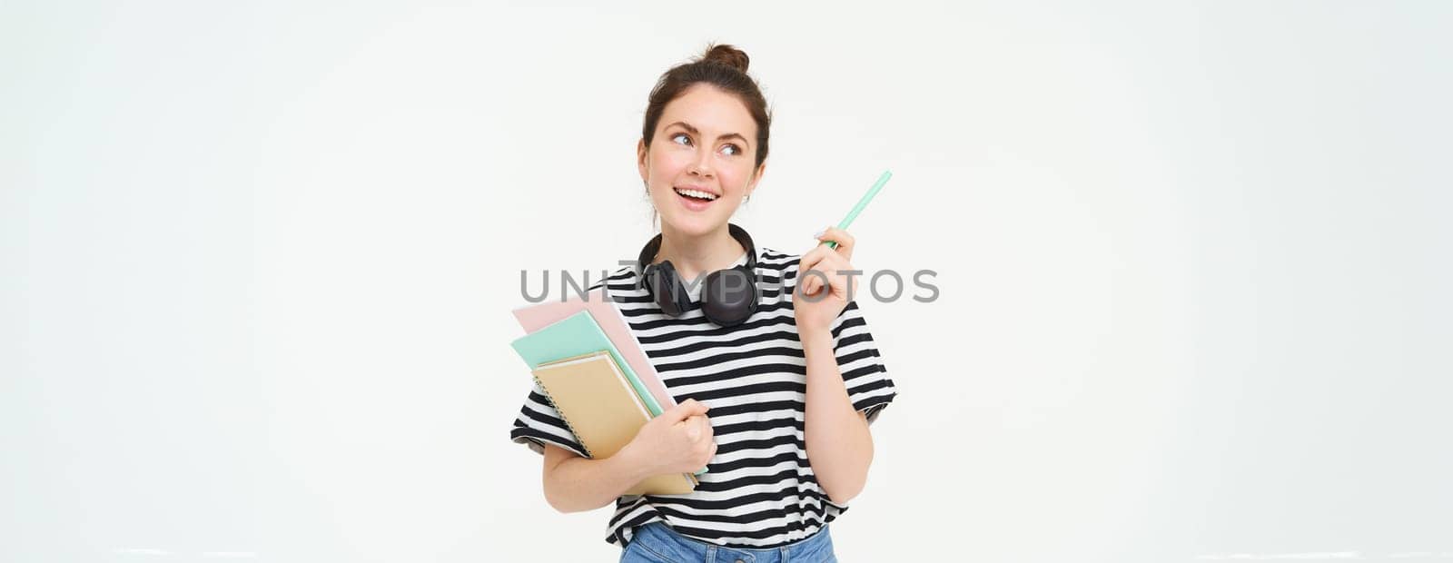 Portrait of young woman, student with notebooks and earphones on her neck, posing for college advertisement, white background.