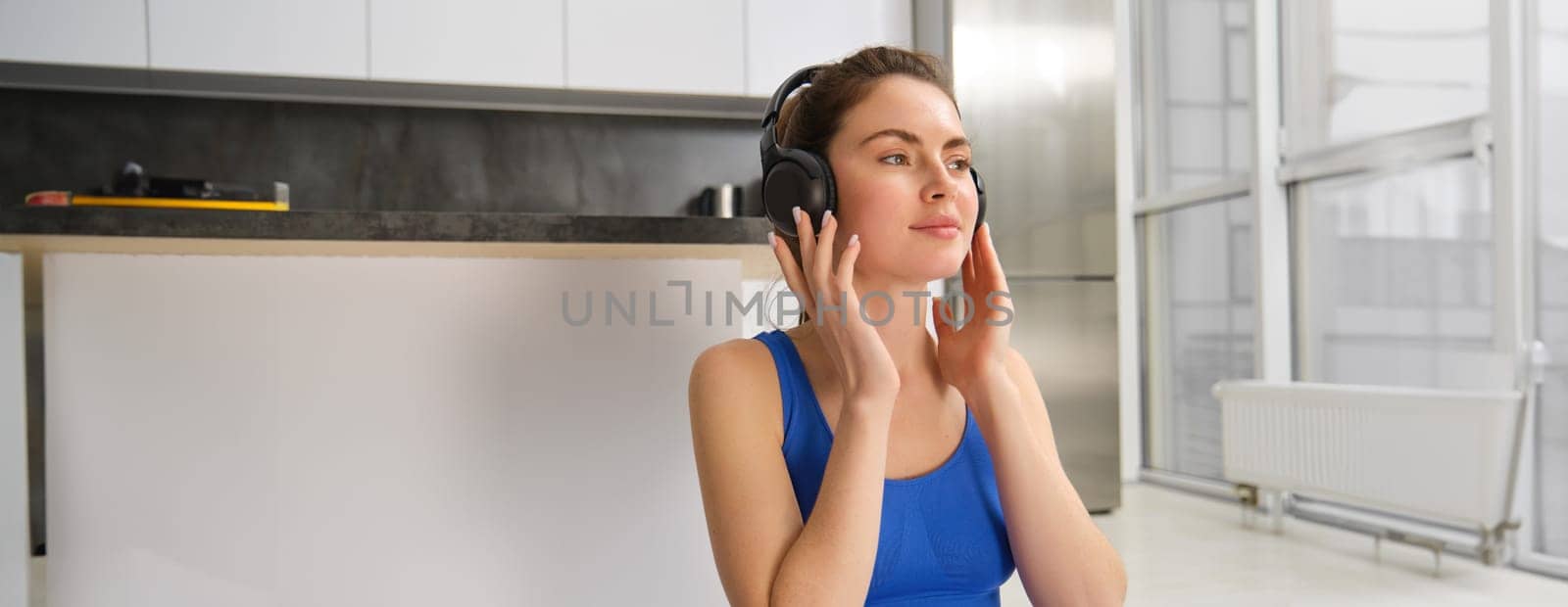Young woman puts on wireless headphones to listen music during workout at home, wears sportsbra, smiles with pleased face.