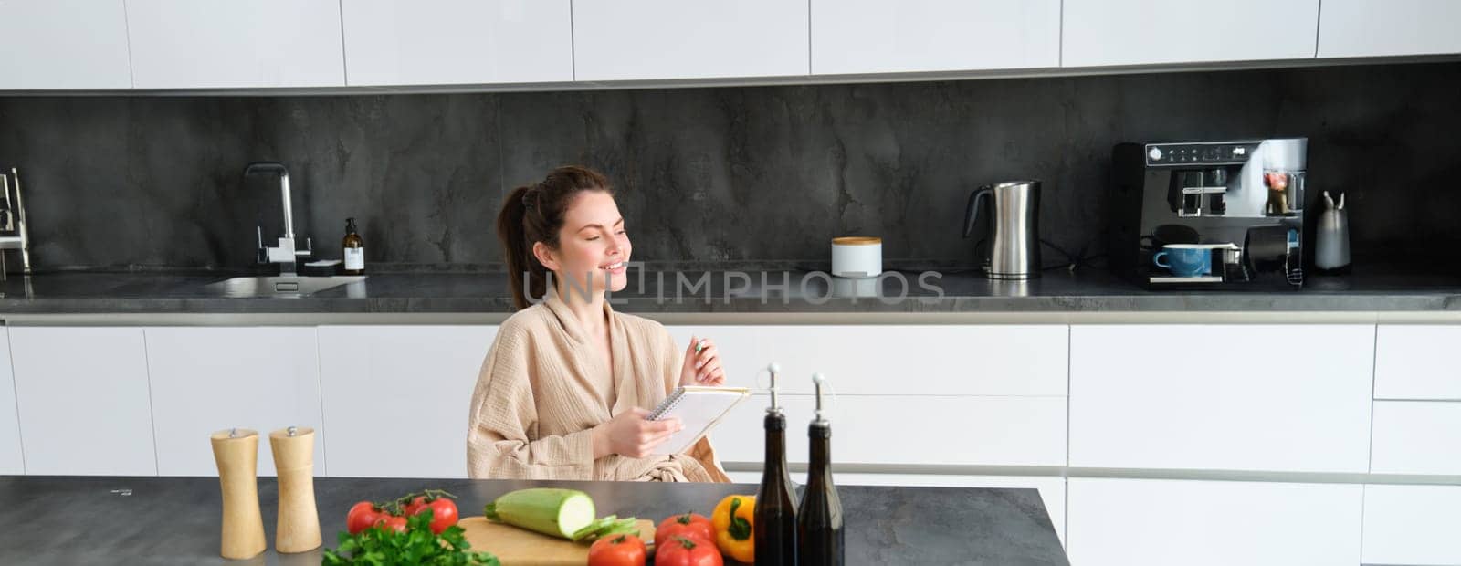 Portrait of woman thinking of menu, sitting in the kitchen and making grocery list, vegetables and meal ingredients on counter by Benzoix