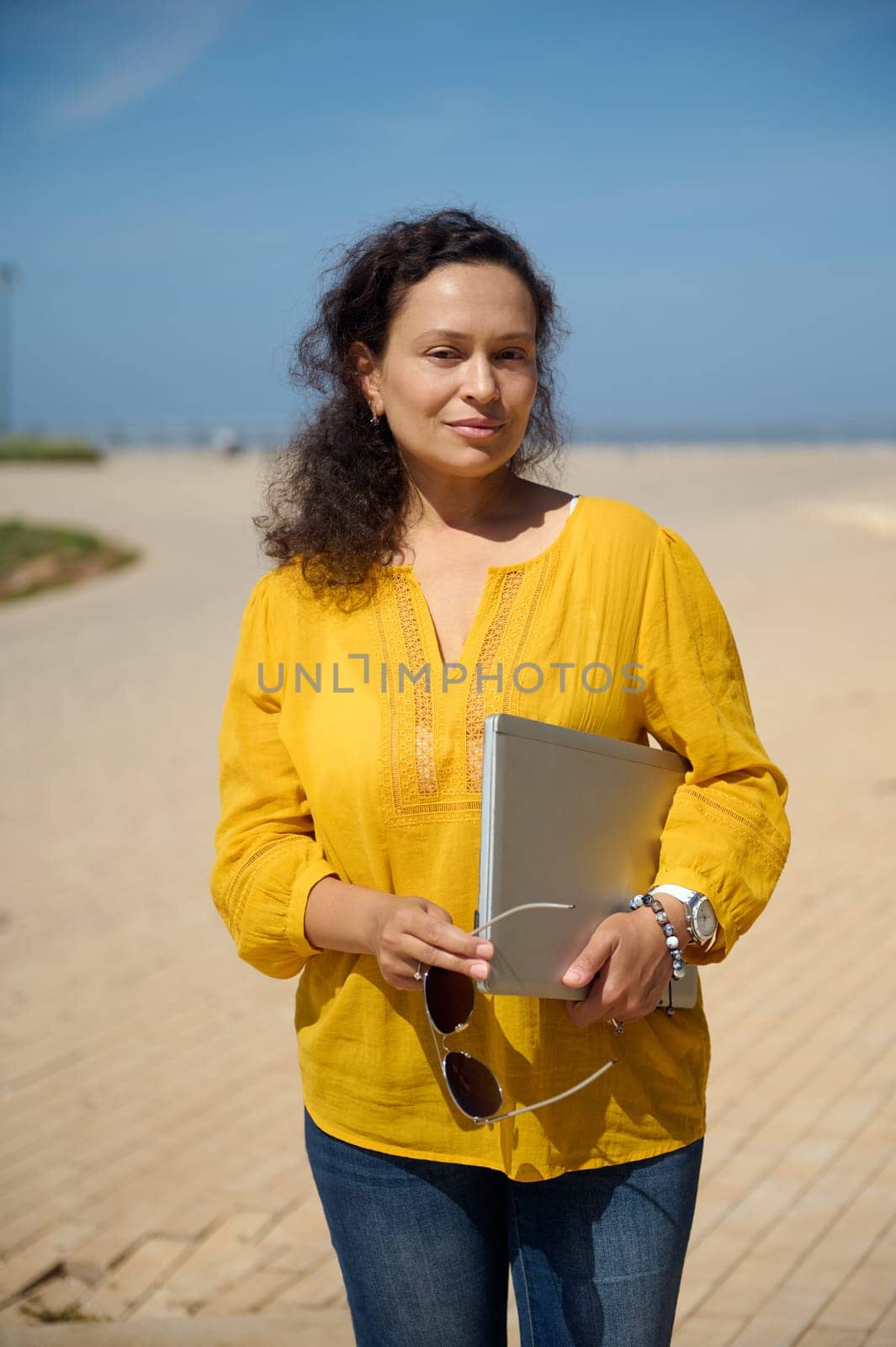 Confident portrait of a beautiful multi-ethnic woman with laptop, looking at camera, standing in the square, dressed in casual yellow shirt and blue denim jeans. Business. People. Recruitment. Career