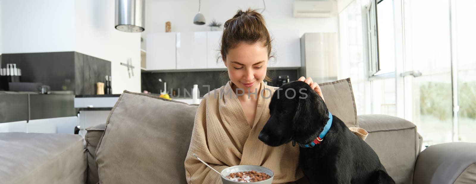 Woman eating cereals with dog on sofa. Breakfast and lifestyle concept