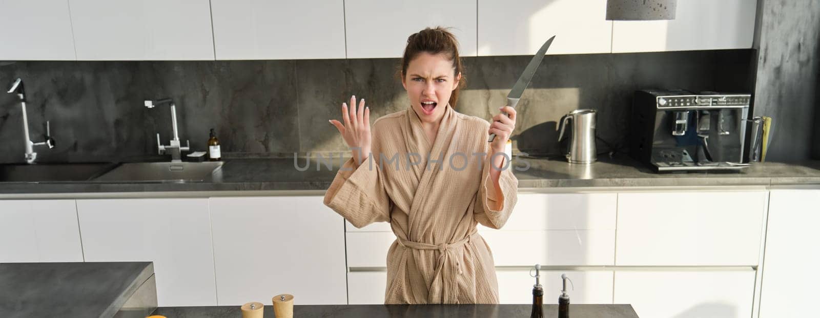 Portrait of annoyed woman with knife, angry while cooking in the kitchen, frustrated while doing house chores and preparing food for family,standing in bathrobe.