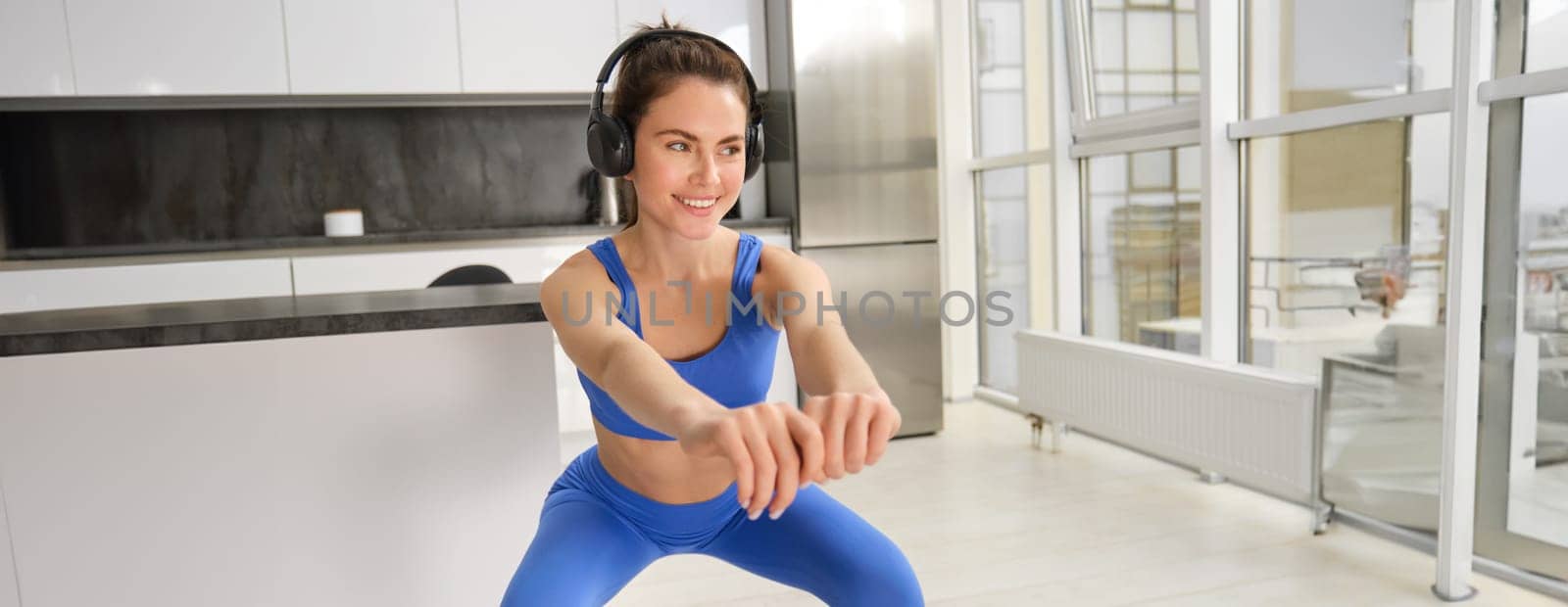 Image of young sportswoman, fitness instuctor doing squats workout at home, listening music in wireless headphones and stretching arms, doing exercises in living room.