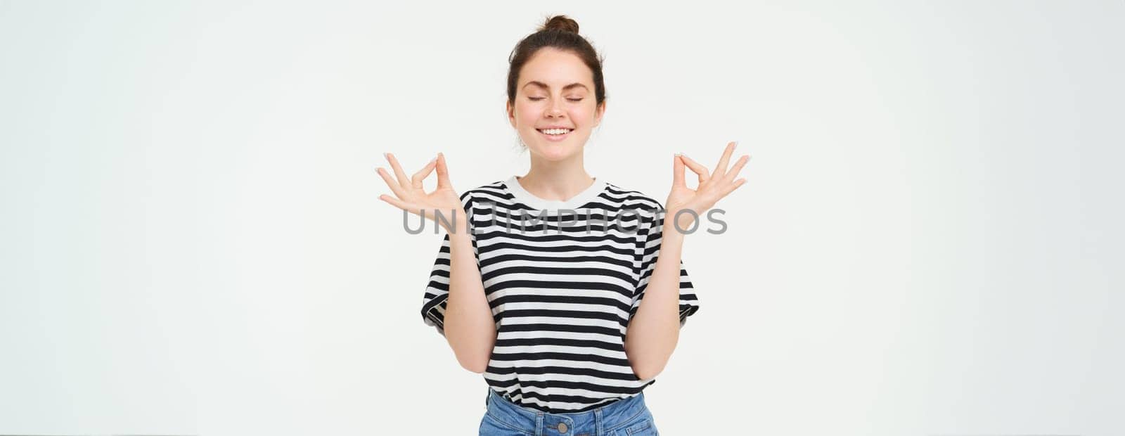 Portrait of young woman relaxing, feeling zen, meditating, smiling with pleased, calm face expression, standing over white background.