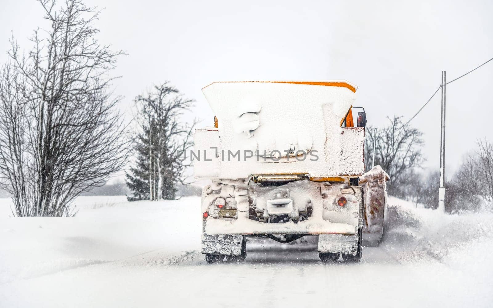 Orange plough truck on snow covered road; gray sky and trees in background; view from car driving behind - winter road maintenance by Ivanko