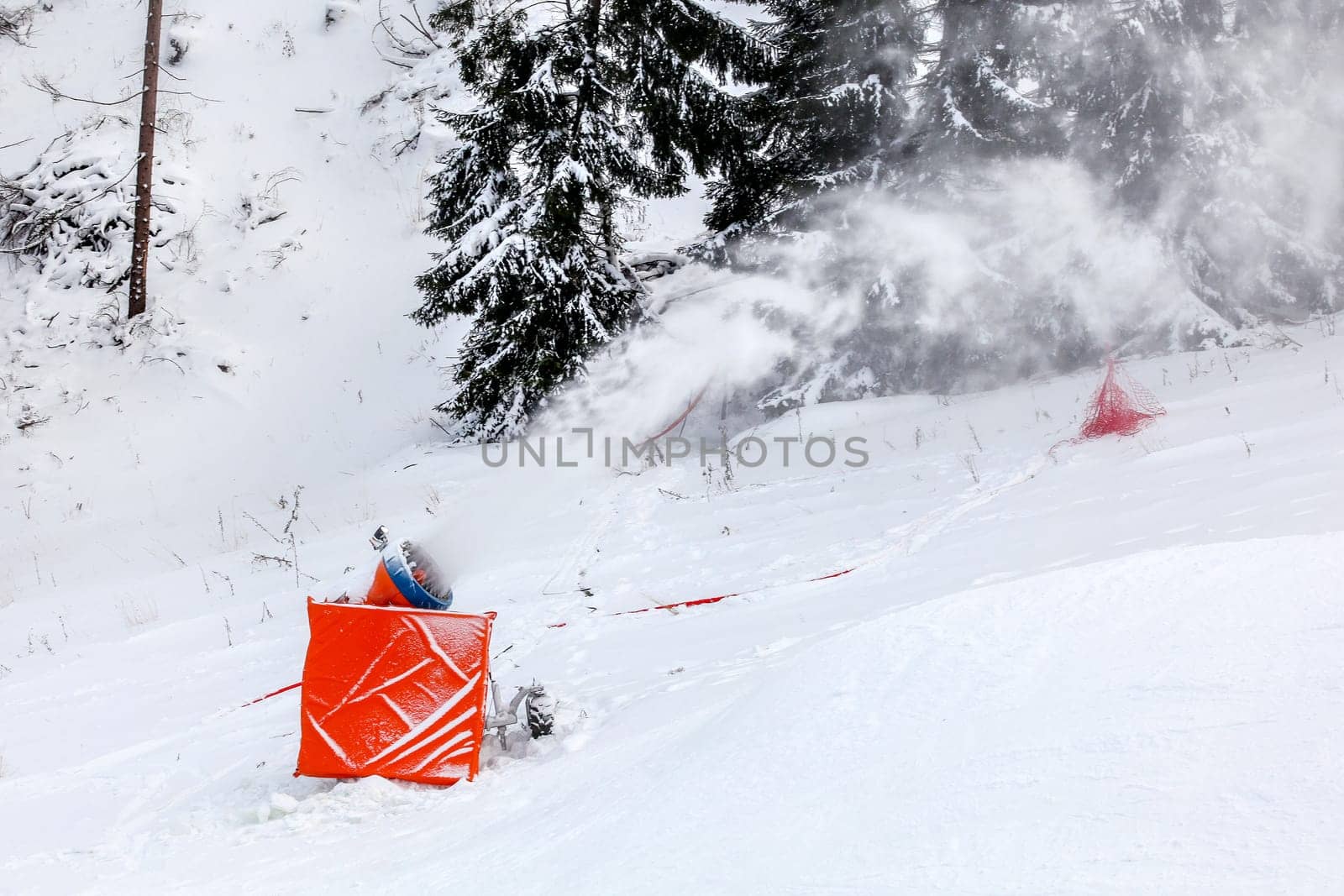 Snow making cannon spraying artificial ice crystals over ski piste, trees in background