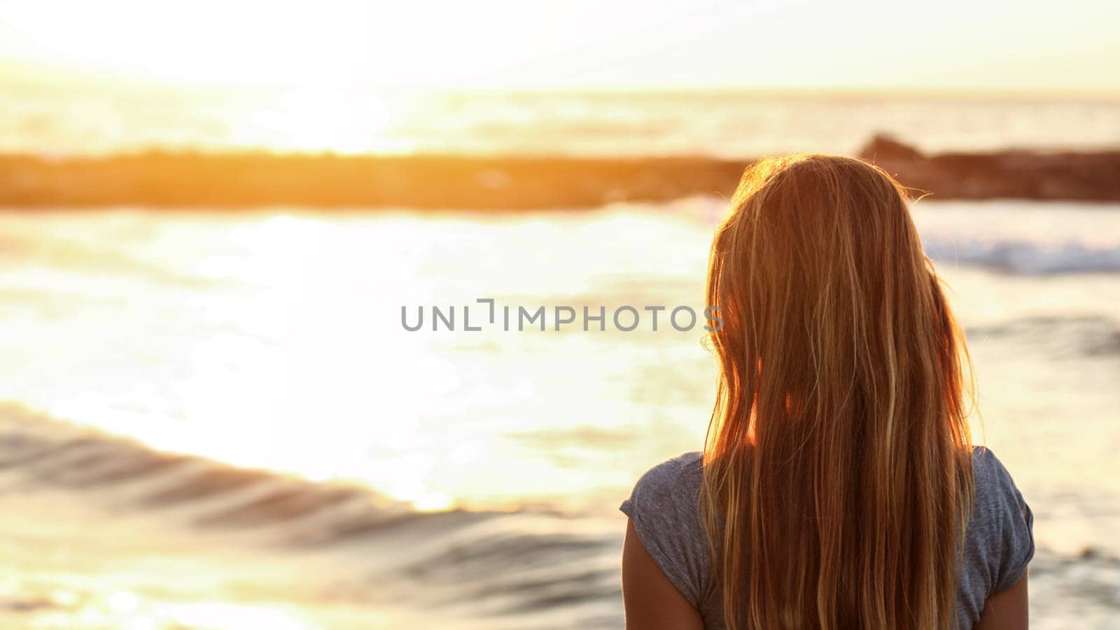 Young woman watches sunset over sea at the beach, view from back, detail on her hair, wide banner with space for text left by Ivanko