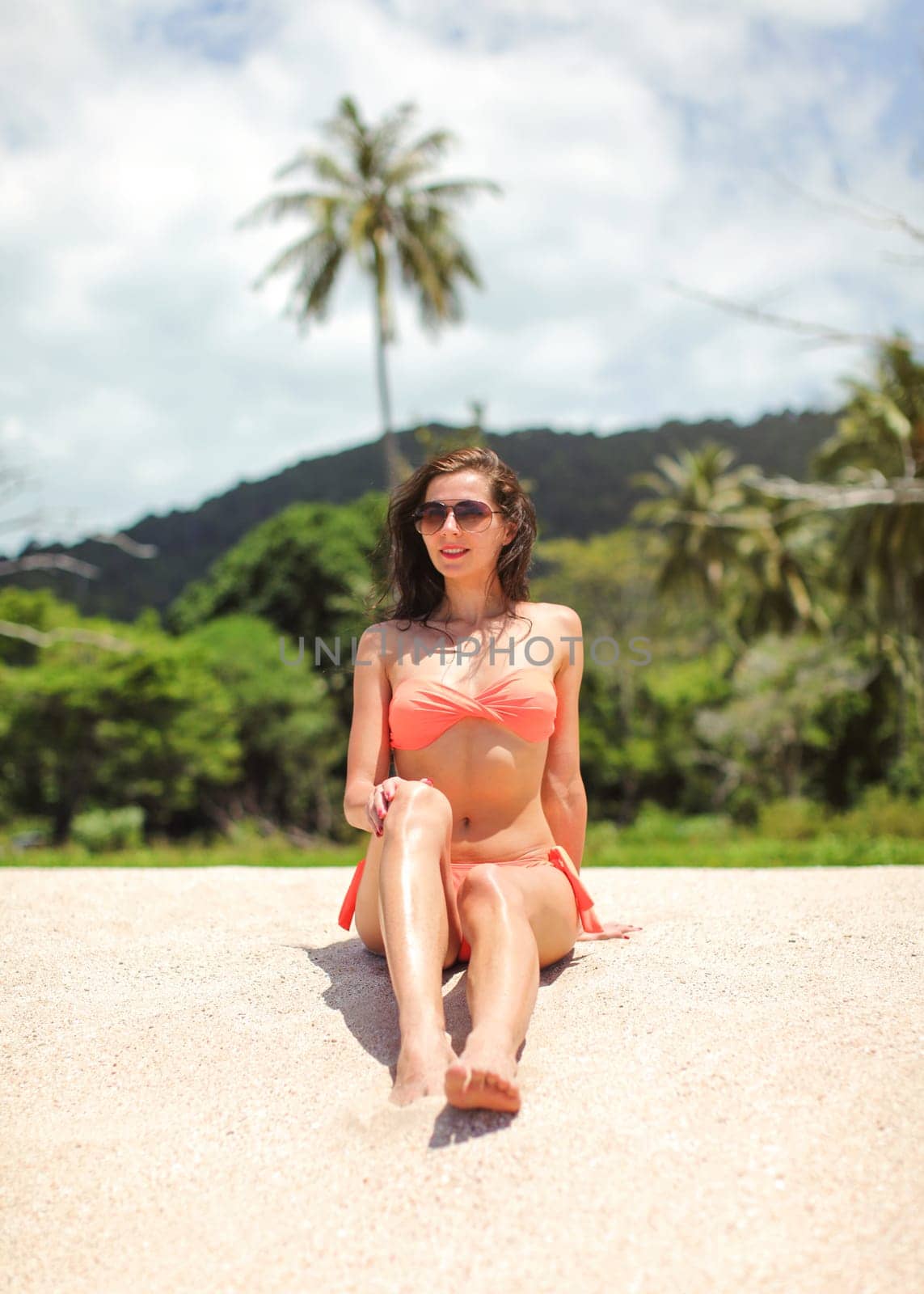 Young slim woman in bikini and sunglasses sitting on fine sand beach, jungle with palm trees behind her.