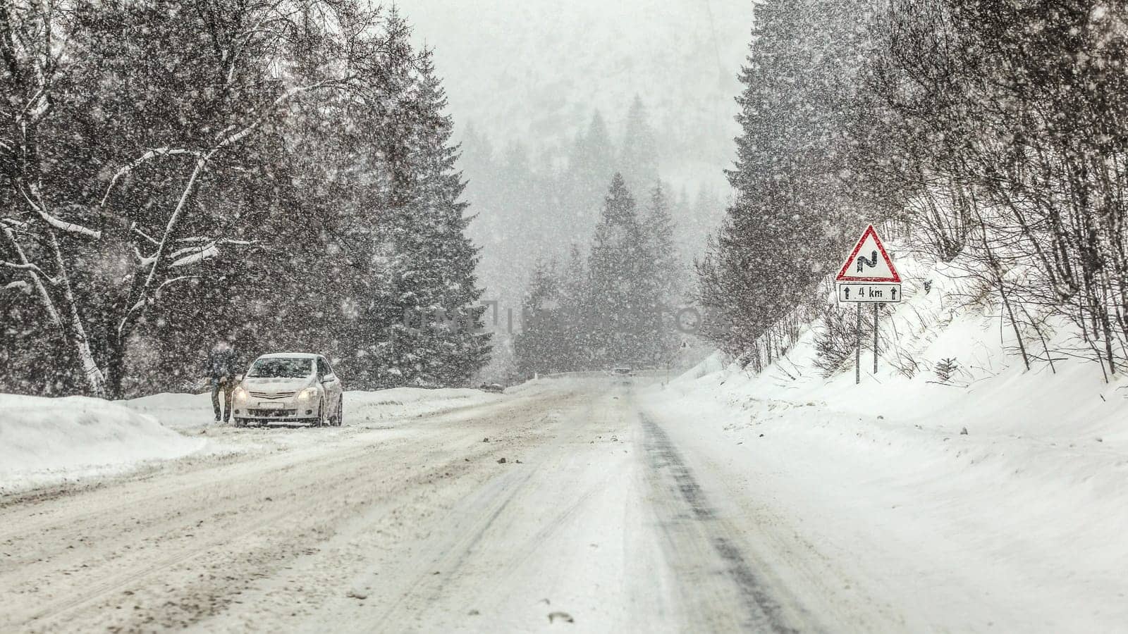 Driving through heavy snowstorm blizzard at forest road, warning curves ahead sign right, and anonymous man with his car stopped on left side of road. View from vehicle driving behind