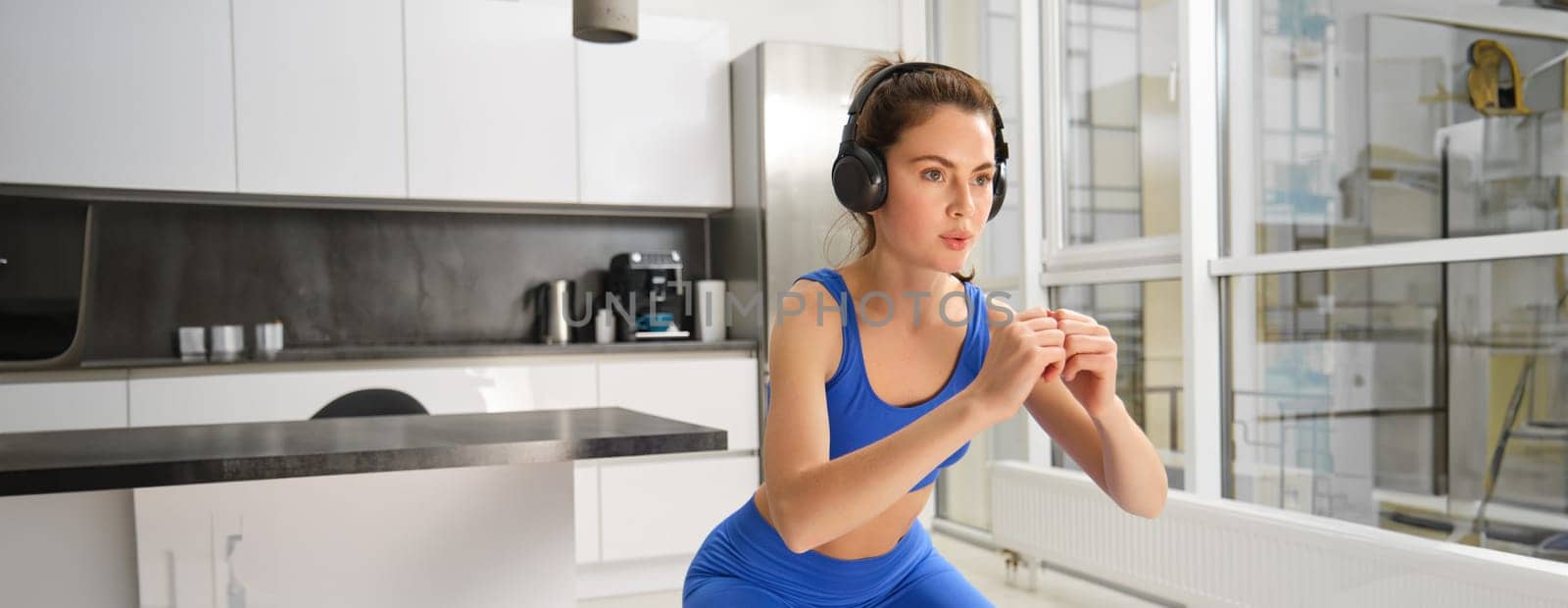 Portrait of woman doing squats with elastic resistance band, looking focused during workout, doing exercises at home.