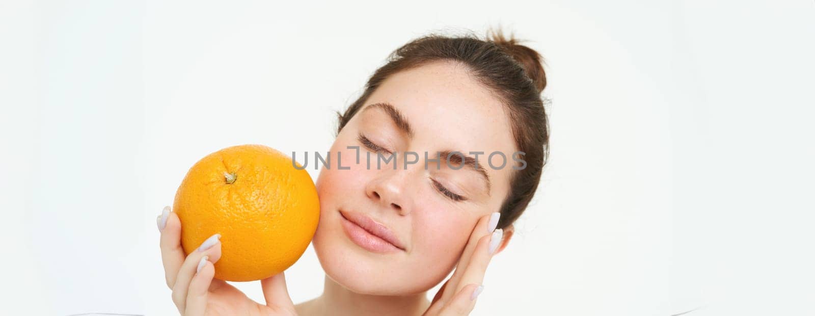 Portrait of young woman holding orange near face without blemishes, smiling, concept of vitamins, nourishment and organic facial treatments, white background.