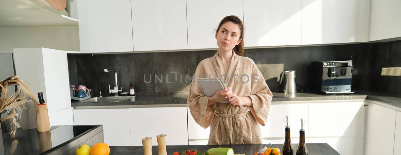 Portrait of creative young woman, cooking, holding recipe book, thinking while making meal, preparing food in the kitchen, standing in bathrobe.