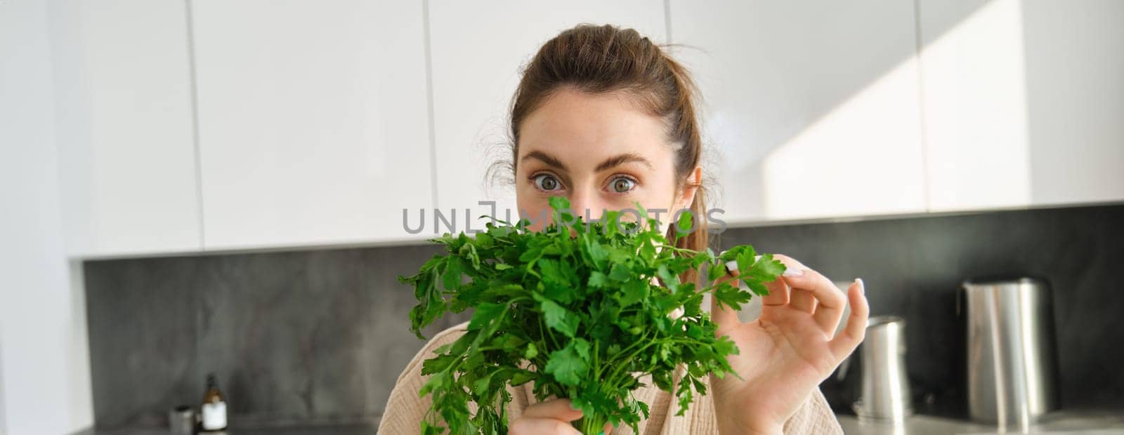 Portrait of beautiful woman cooking, holding fresh parsley, adding herbs while making salad, healthy meal in the kitchen.