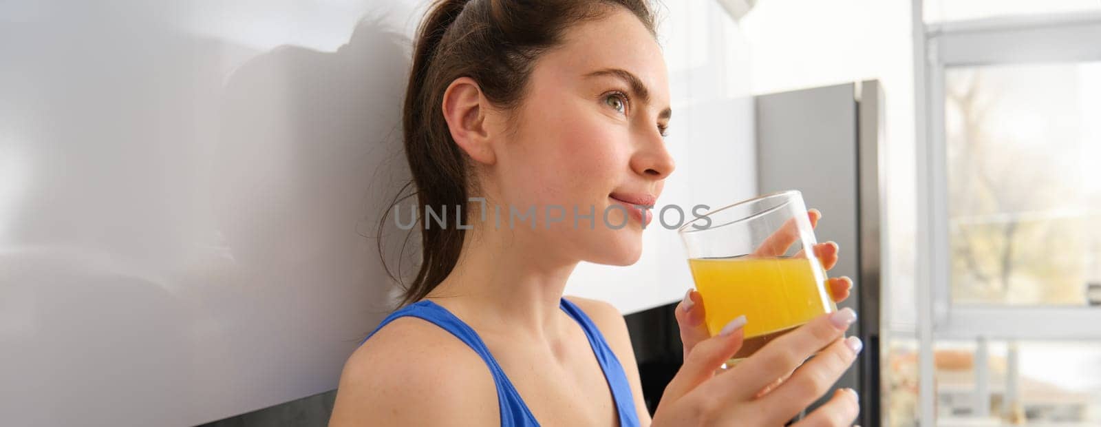 Close up portrait of beautiful young woman, looking away, drinking orange juice, fresh in glass, standing in kitchen, wearing workout clothes.