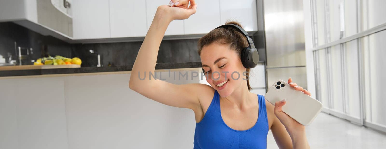 Portrait of happy, brunette woman in headphones, holding smartphone, dancing and working out, doing fitness exercises on yoga mat.