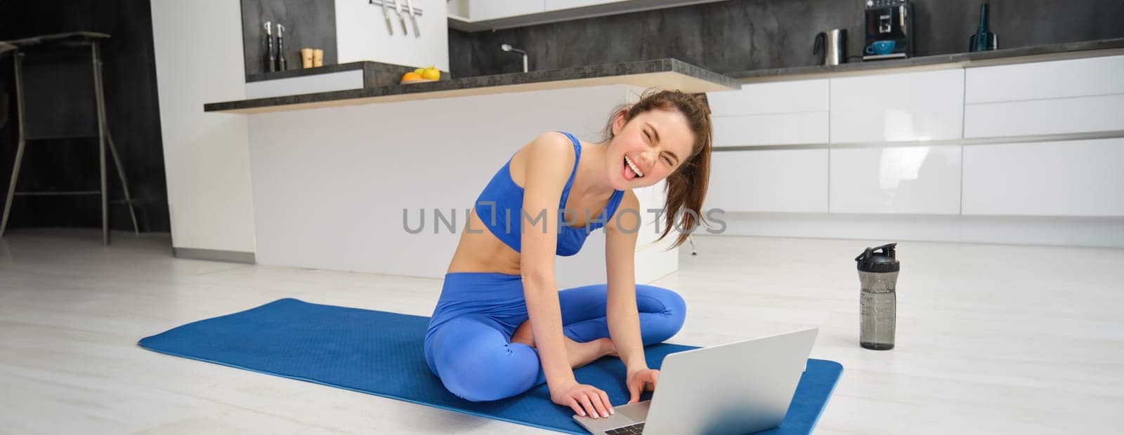 Portrait of happy young fitness woman, talking with client over laptop video chat, records her workout training session, showing aerobics exercises.