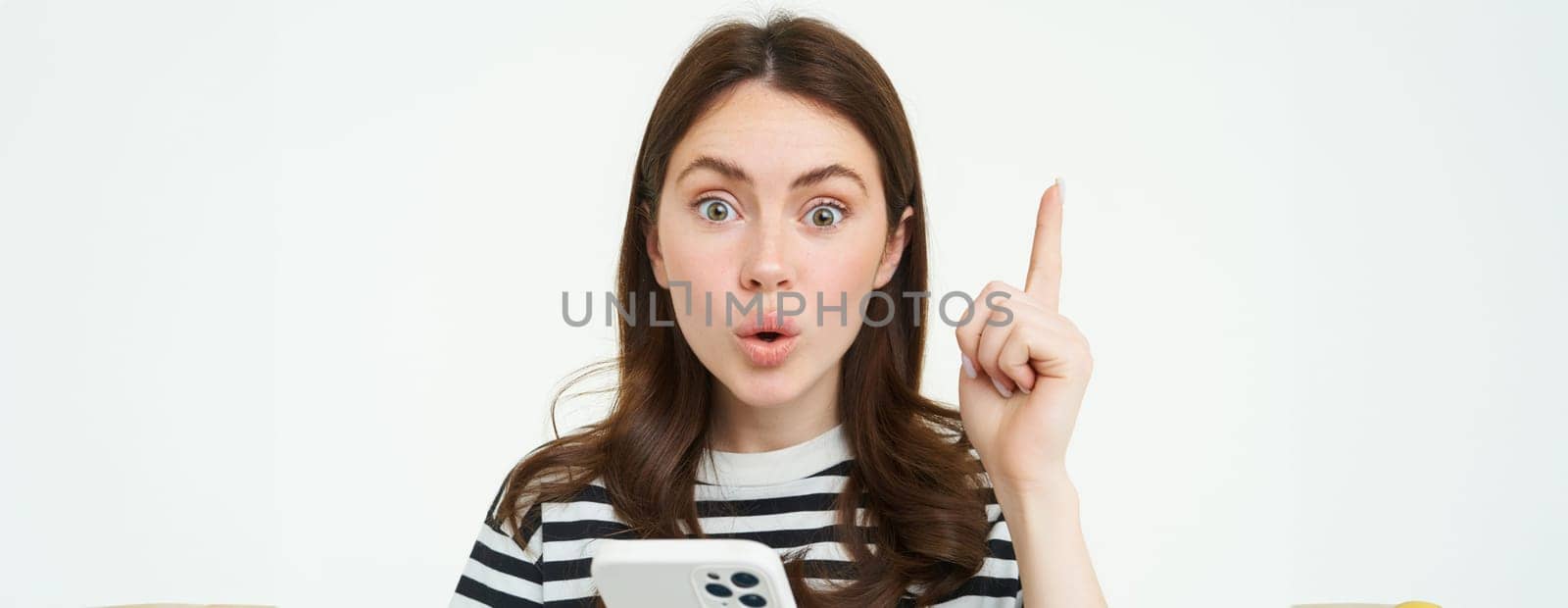 Portrait of excited woman raising finger, eureka gesture, has an idea or solution, holds mobile phone, white background.